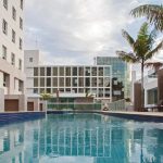 No lado esquerdo, uma parte do edifício do Jade Hotel Home Office com destaque para a faixa de vidro verde. No meio até a extremidade direita, destaque para a piscina de cor azul e ao fundo, a vista do prédio comercial horizontal do Jade Hotel, com destaque para a vidraça verde. No fundo, ao lado direto, há dois coqueiros compondo a imagem.